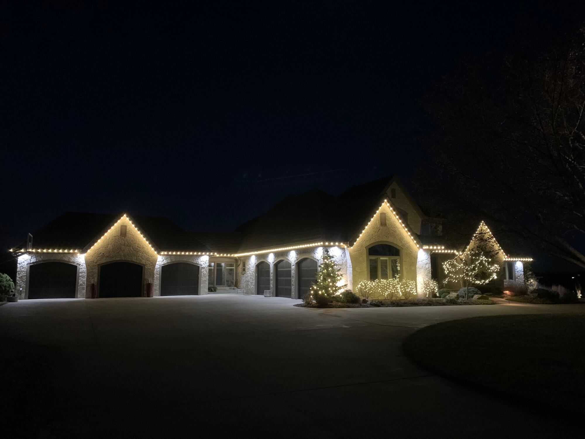 Warm white roofline lighting on a white stone home with lit evergreens at the entry